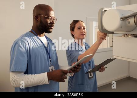 Due giovani medici in uniforme accendono le apparecchiature mediche in ambiente ospedaliero. La scena mette in evidenza il lavoro di squadra e l'assistenza sanitaria moderna in azione. Adatto per ospedale, clinica e campagne di marketing di assistenza sanitaria. Foto Stock