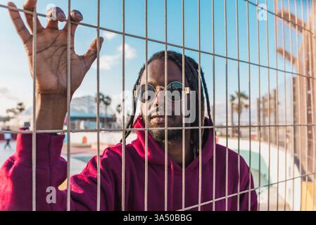 Un uomo nero serio con dreadlock che indossano occhiali da sole e una felpa con cappuccio si trova dietro una recinzione di metallo in un parco in una giornata di sole. Il look urban Street style abbina un abbigliamento casual a un mood attento, ideale per lo stile di vita e le campagne urbane. Foto Stock