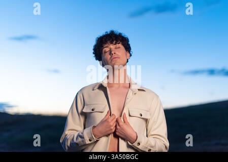 L'uomo ispanico con i capelli ricci sta all'aperto al crepuscolo con gli occhi chiusi, tenendo la sua camicia. L'umore calmo e riflessivo si adatta alla consapevolezza, al relax o alla grafica dello stile di vita. Una leggera luce al crepuscolo migliora il ritratto. Foto Stock