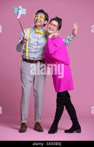 Allegra coppia cinese senior che si fa un selfie con uno smartphone. Questa immagine calda e tecnologica è ideale per campagne di invecchiamento, viaggi o lifestyle. Lo spazio di copia sul lato consente di inserire il testo. Foto Stock