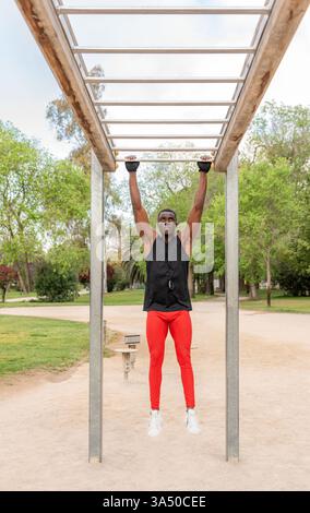 Serio giovane atleta afro-americano maschile in abbigliamento sportivo che fanno tira in su esercizio sul bar durante l'allenamento in strada Foto Stock