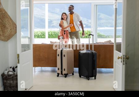 Coppia felice e variegata che abbraccia nel soggiorno con valigie, godendo dell'arrivo in vacanza Foto Stock