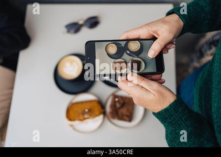 Donna ispanica che scatta foto di caffè e dolci sul suo smartphone mentre siede al tavolo in un caffè all'aperto durante il giorno Foto Stock