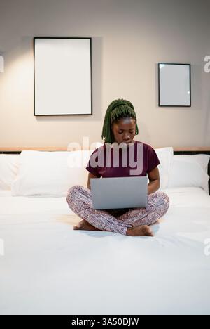Una donna nera seria siede a gambe incrociate su un letto, lavorando su un notebook in una camera da letto di casa. La scena enfatizza il telelavoro e la messa a fuoco con un'atmosfera pulita a casa. Ideale per il lavoro, il freelance o il lavoro da remoto, con un ambiente domestico professionale. Foto Stock