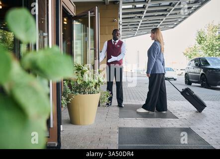 Il fattorino nero dà il benvenuto a una donna caucasica con bagagli fuori dall'ingresso dell'hotel durante il giorno. Un momento di ospitalità amichevole, perfetto per viaggi e alloggio. Ottimo per la reception, i servizi per gli ospiti e le campagne turistiche. Foto Stock