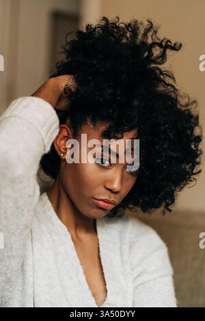 Tranquillo ritratto di una donna nera che si rilassa a casa. Si siede su un divano, toccando i capelli in un salotto tranquillo e confortevole. Questa immagine si adatta bene allo stile di vita e alle caratteristiche della vita domestica, mettendo in risalto lo stile degli interni e il benessere personale. Foto Stock