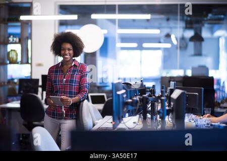 Donna d'affari nera sorridente con Afro che tiene in mano un tablet in un moderno ambiente d'ufficio. Immagini professionali per lo stile di vita aziendale, ideali per campagne tecnologiche, di leadership e sul luogo di lavoro. Adatto per progetti a tema business, imprenditorialità e successo. Foto Stock