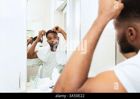 Foto di stile di vita informale di un giovane uomo nero che si controlla i capelli nello specchio del bagno. L'impostazione luminosa e riflettente cattura i momenti di cura personale e di cura del corpo, con ampio spazio per la copia. Ideale per campagne di bellezza, cura del corpo o benessere rivolte agli uomini. Foto Stock