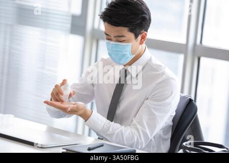 Uomo d'affari cinese che indossa la maschera usando igienizzatore per le mani seduto alla sua scrivania con il computer portatile in ufficio Foto Stock