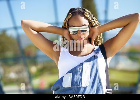 Donna nera casual ed elegante che guarda la fotocamera e indossa occhiali da sole moderni. Questa immagine di stile Street-style si adatta alla moda, alla vita urbana e al marchio di fiducia. Foto Stock