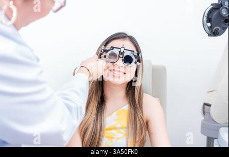 Primo piano di un paziente sorridente durante un esame oculistico con un optometrista utilizzando un casco di prova. L'immagine mette in evidenza la cura degli occhi, i test visivi e la professionalità clinica. Adatto per argomenti di sanità e optometria con spazio di testo. Foto Stock