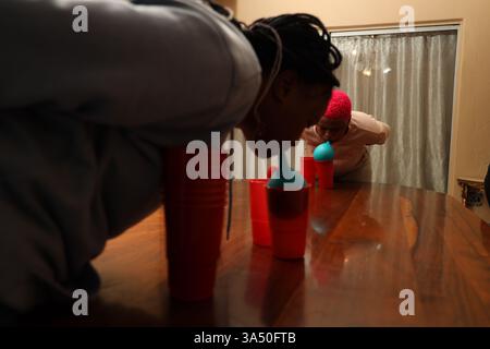 Le sorelle adolescenti nere focalizzate in casa, in piedi con le mani dietro la schiena mentre impilano le tazze su un tavolo. Questo momento divertente cattura il legame tra fratelli e colleghi e il lavoro di squadra, ideale per le campagne in famiglia e per lo stile di vita. Ideale per interni, soggiorno e attività domestiche. Foto Stock
