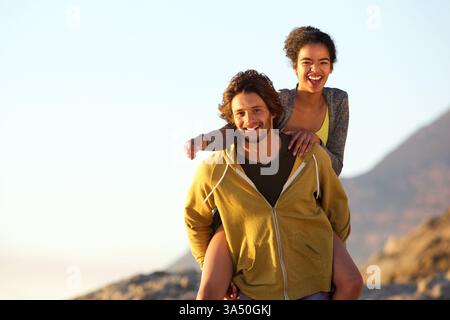 Allegra coppia multirazziale all'aperto al tramonto, con la donna che fa il piggyback sul suo partner. Il momento irradia calore, giocosità e insieme. Perfetto per lo stile di vita, gli appuntamenti e le immagini di viaggio. Foto Stock