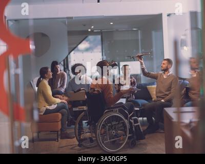 Un uomo d'affari disabile in sedia a rotelle partecipa a una riunione di gruppo in un moderno ufficio di coworking open space. Si occupa di occhiali di protezione per la realtà virtuale e di assistenza con i droni, evidenziando l'accessibilità e l'innovazione. Un'immagine ideale per la tecnologia inclusiva, la cultura delle startup e i luoghi di lavoro futuri. Foto Stock