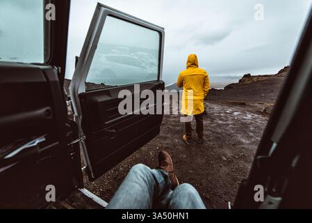 esploratore maschio seduto in auto vicino a un amico maschio che indossa una giacca gialla in piedi e guarda il panorama mentre viaggia attraverso l'Islanda alla scoperta del nat... Foto Stock