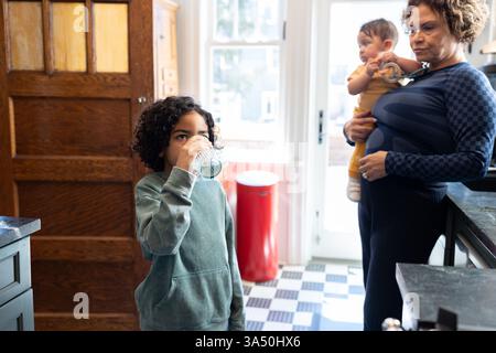 Donna ispanica di mezza età con capelli ricci sta in cucina con la nipote multirazziale, mentre guarda suo nipote bere acqua. Un caldo momento familiare in casa che mette in risalto la vita di tutti i giorni e il legame intergenerazionale. Ideale per la vita a casa, i genitori e le immagini dello stile di vita familiare. Foto Stock