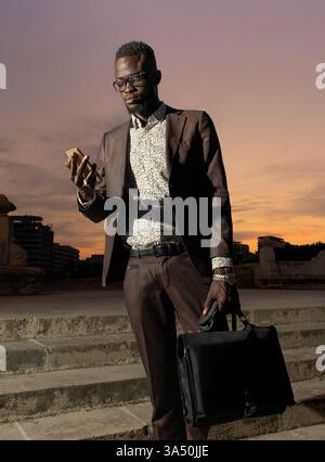Giovane imprenditore afroamericano concentrato in una tuta elegante si stende con una valigetta, che utilizza uno smartphone su un caldo sfondo urbano. Questa immagine si adatta ai temi aziendali e tecnologici, evidenziando connettività e professionalità. Foto Stock