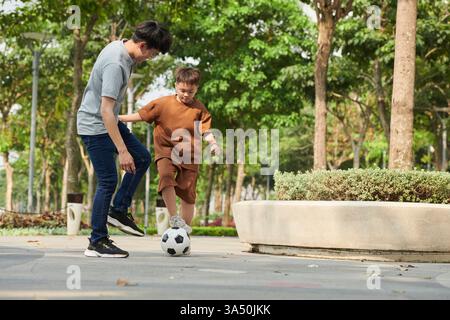 Padre e figlio giocano a calcio in un parco la mattina del fine settimana, catturando la famiglia che gioca all'aperto. Adatto per attività in famiglia, sport e immagini per il tempo libero del fine settimana. Foto Stock