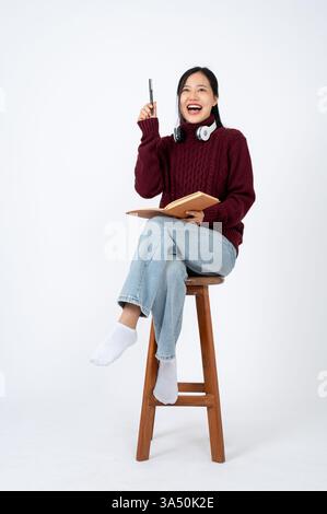 Una giovane donna asiatica siede con un diario, pensando e progettando idee in un luminoso studio. L'umore riflettente si adatta alle campagne creative, educative e lifestyle, con opportunità di spazio di copia e temi giornalistici. Foto Stock
