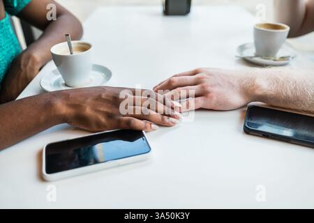 Coppia multirazziale seduta in un bar diurno, mani che toccano accanto alle tazze da caffè e uno smartphone. Ideale per campagne di lifestyle, incontri e relazioni con un'atmosfera calda e contemporanea. La scena di un caffè candid con luce naturale migliora il romanticismo e la connessione. Foto Stock