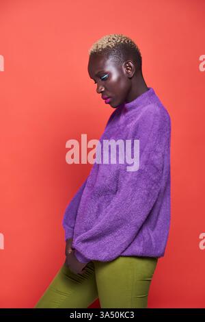 Vista laterale del ritratto di un'elegante donna africana con i capelli corti in uno studio su uno sfondo rosso audace. L'illuminazione da studio mette in risalto una presenza sicura e alla moda. Adatto per campagne di ritratto, stile e cosmetici. Foto Stock