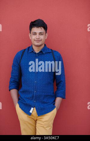 Ritratto di un uomo d'affari indiano informale con una camicia blu e uno zaino contro una parete rosa. Ideale per immagini di avvio e imprenditorialità con un tocco di colore contemporaneo. La scena comunica un marchio moderno e sicuro e la prontezza professionale. Foto Stock