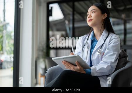 Professionista asiatico donna medico siede nel suo ufficio, lavorando su un tablet digitale e rivedere i casi medici. Questa immagine sanitaria trasmette competenza e moderna pratica medica. Adatto per comunicazioni mediche, sanitarie e cliniche. Foto Stock