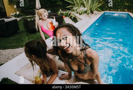 Sorridente donna asiatica appoggiata sulla piscina durante una vivace festa in piscina con gli amici. Perfetto per viaggi, vacanze estive e grafica stile resort. Foto Stock
