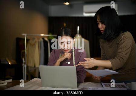 Le donne stilisti asiatiche collaborano in un'affollata sartoria, rivedendo i tessuti e discutendo di disegni di abiti. Il team creativo dimostra il lavoro di squadra in uno studio di moda, con la moda sartoriale e boutique in primo piano. Ideale per articoli su studi di moda di startup, designer millennial e progetti di design collaborativi. Foto Stock