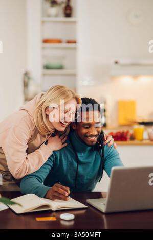 La moglie bionda mostra sostegno mentre suo marito lavora da casa su un laptop. La coppia si siede insieme in uno spazio abitativo moderno, illustrando il lavoro remoto e la collaborazione. Ideale per la vita familiare, le campagne di lavoro da casa e le immagini di stili di vita multiculturali. Foto Stock