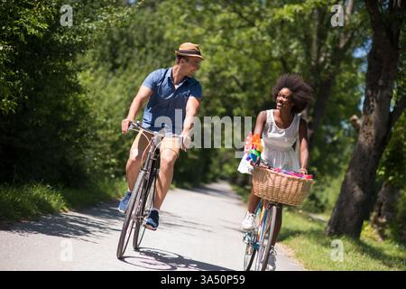 Giovani coppie in bicicletta insieme in un parco in una soleggiata giornata estiva, sorridendo e godendo il momento. Questa immagine funziona bene per lo stile di vita, gli incontri, la salute e i temi di ricreazione all'aperto. Foto Stock