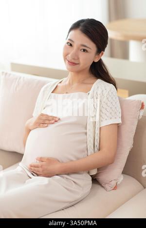 Felice donna cinese incinta che si rilassa su un divano a casa con un comodo abbigliamento per la maternità. Questa immagine di stile di vita gioioso si adatta alle campagne sulla gravidanza, la famiglia e il comfort domestico. Foto Stock