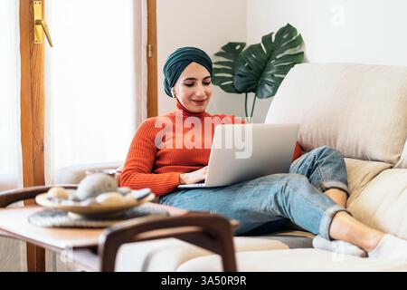 Una donna musulmana con un elegante velo sorride mentre utilizza un laptop su un divano a casa, ascoltando musica nelle giornate di sole. La scena domestica e rilassata si adatta allo stile di vita, al lavoro da remoto e al branding della cultura digitale. Ideale per l'ufficio domestico, l'apprendimento online e la messaggistica positiva. Foto Stock