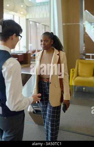 Donna d'affari di colore con bagagli in conversazione con un receptionist asiatico che tiene un tablet nella hall dell'hotel. Ideale per immagini di ospitalità con lavoro di squadra interculturale e servizi agli ospiti in viaggio. Una scena professionale in un ambiente alberghiero affollato. Foto Stock