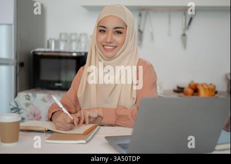 Bella studentessa asiatica musulmana studia a un tavolo in cucina, scrivendo appunti nel suo taccuino. Funziona bene per l'istruzione, la vita studentesca e i temi di apprendimento remoto. Foto Stock