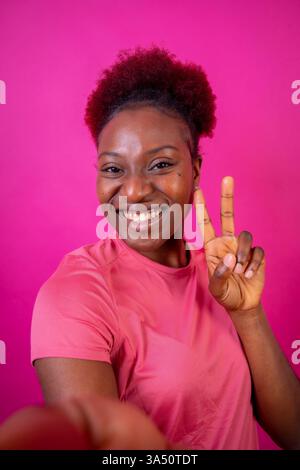 Giovane afroamericana isolata su uno sfondo rosa sorridente con il gesto della vittoria, ripresa in studio Foto Stock