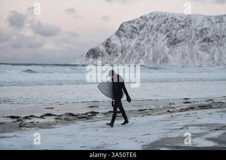 Autentico surfista artico sulla costa norvegese dopo una sessione nei freddi mari del nord. Una straordinaria immagine di sport estremi con surf invernale e aspro paesaggio costiero. Foto Stock