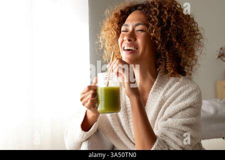 Felice donna multirazziale godendo di sano succo verde a casa. Donna afroamericana che fa disintossicazione bevendo frullato verde. Copia spazio. Uno stile di vita sano Foto Stock