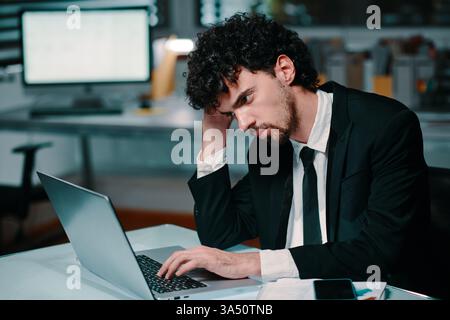 Uomo d'affari di tipo misto che scrive su un notebook di notte in ufficio. La scena comunica concentrazione, produttività notturna e dedizione professionale. Utile per temi quali prestazioni lavorative, scadenze e imprenditorialità aziendale. Foto Stock