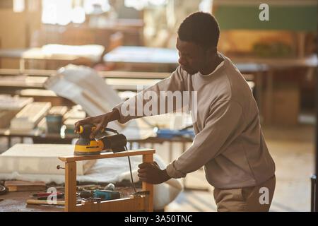 Vista laterale del ritratto di un giovane falegname afroamericano in un negozio di legno illuminato dal sole, che costruisce mobili in legno. Questa immagine mette in risalto le abili falegnamerie in azione con utensili e legno, oltre a un ampio spazio di copia per i layout. Perfetto per caratteristiche di artigianato, mobili fatti a mano e mestieri. Foto Stock