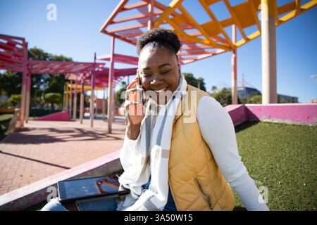 Una allegra donna afroamericana siede in un parco cittadino in una giornata di sole, parlando al telefono. Possiede un tablet, evidenziando uno stile di vita connesso e mobile. Ideale per la vita urbana, la comunicazione digitale e i temi del lavoro da remoto. Foto Stock