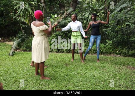 Vibrante ritratto all'aperto di una donna nera con un afro corto rosa che scatta una foto di amici in un parco durante il giorno. Il gruppo gode di sole, vegetazione e un'atmosfera spensierata e amichevole. Ideale per lo stile di vita, l'amicizia e le attività all'aperto. Foto Stock