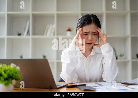 Una donna d'affari asiatica stressata siede alla scrivania del suo ufficio, visibilmente preoccupata per una scadenza imminente. La scena trasmette la pressione sul luogo di lavoro e il lavoro dedicato in un ambiente d'ufficio moderno. Foto Stock