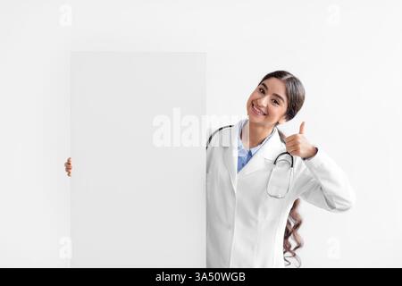 Il milionario medico indiano sorridente in un cappotto bianco regge un grande striscione di carta con spazio vuoto e regala un pollice in alto. Ideale per promozioni sanitarie e pubblicità predisposta per la copia. Foto Stock