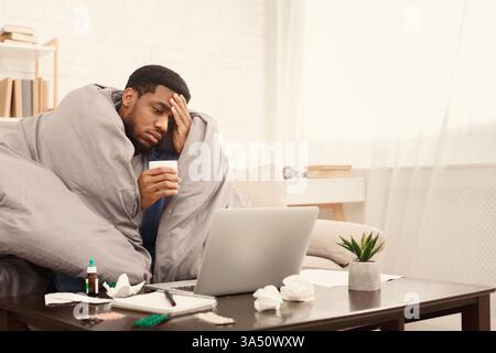 Un dipendente afroamericano malato lavora da casa, prendendo pillole influenzali mentre si utilizza un computer portatile, con spazio di copia per la messaggistica. Si adatta agli articoli sul lavoro a distanza, sulle malattie sul posto di lavoro e sulla continuità aziendale. Foto Stock