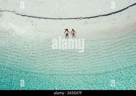 Donna asiatica e uomo caucasico che prendono il sole insieme su una spiaggia di sabbia bianca su un'isola privata. Questa destinazione tropicale trasmette svago, viaggi di coppia e fughe romantiche in un ambiente incontaminato. Perfetto per viaggi, stile di vita e destinazioni con sole, mare e vacanze sull'isola. Foto Stock