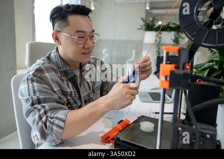 L'uomo asiatico in un ufficio moderno progetta parti meccaniche utilizzando una stampante 3D. Questa immagine di ingegneria e tecnologia è ideale per gli argomenti STEM, innovazione e produzione. Ideale per articoli su progettazione, prototipazione e fabbricazione digitale. Foto Stock