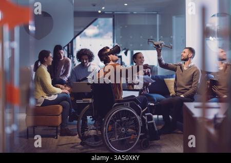Un uomo d'affari disabile in sedia a rotelle partecipa a una riunione di gruppo in un moderno ufficio di coworking open space, utilizzando occhiali di protezione per la realtà virtuale e l'assistenza dei droni. La scena mette in evidenza diversità, accessibilità e collaborazione tecnologica. Ideale per articoli su luoghi di lavoro inclusivi, startup e innovazione. Foto Stock