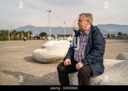 Uomo anziano in giacca casual seduto sulla panca di pietra, godendo dell'aria fresca e della natura. Concetto di pensione, vita tranquilla, relax all'aperto e benessere degli anziani. Foto Stock