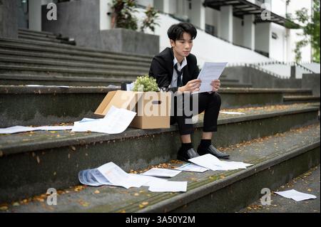 Un giovane uomo d'affari asiatico, stressato e depresso, sta leggendo la sua lettera di licenziamento mentre si siede sulle scale davanti all'ufficio aziendale con un Foto Stock